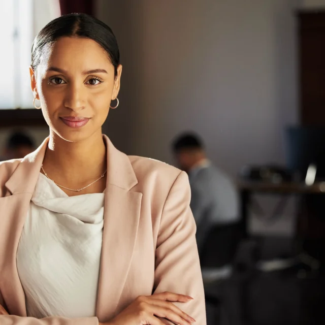 A woman in professional attire stands with arms crossed, looking at the camera, in an office setting with two people working in the background.