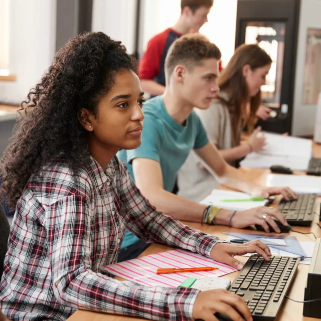 Students sitting at desks work on desktop computers in a classroom setting, with notebooks and stationery on their desks.