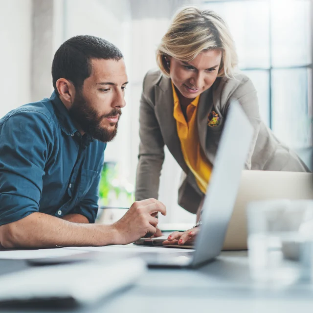Two colleagues in an office review information on a laptop together, with documents and a pen on the desk in front of them.