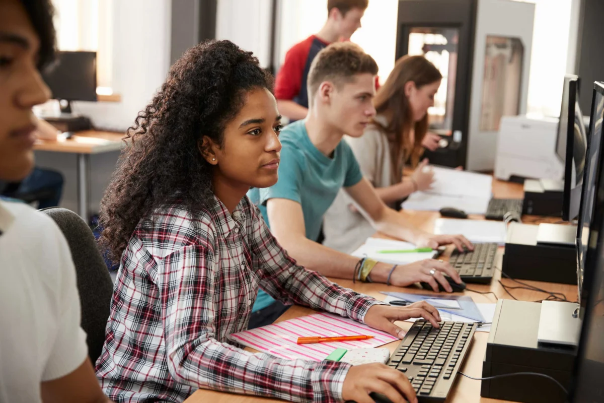 Several students sit at desks using desktop computers in a classroom, focusing on their work. Notebooks, pens, and papers are scattered on the desks.