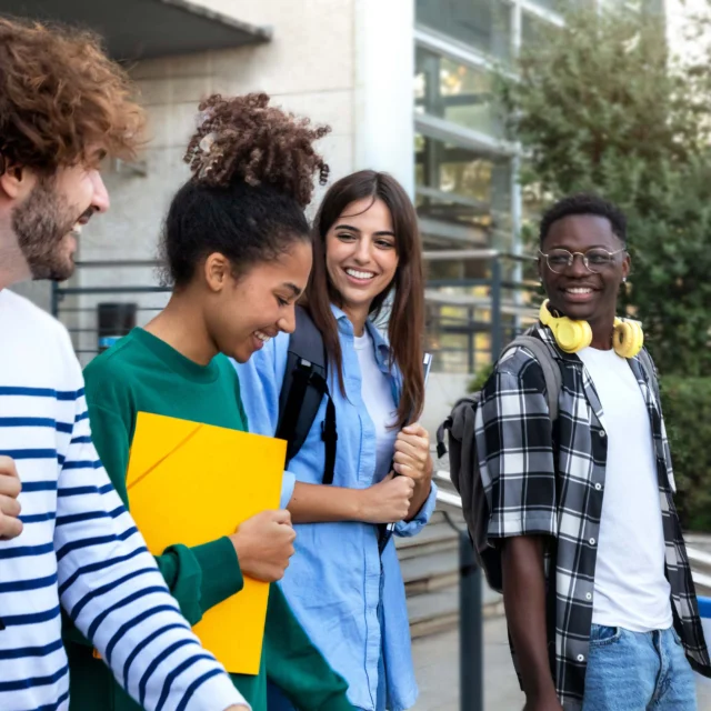 Four students with backpacks and books walk together outside a building, smiling and talking.