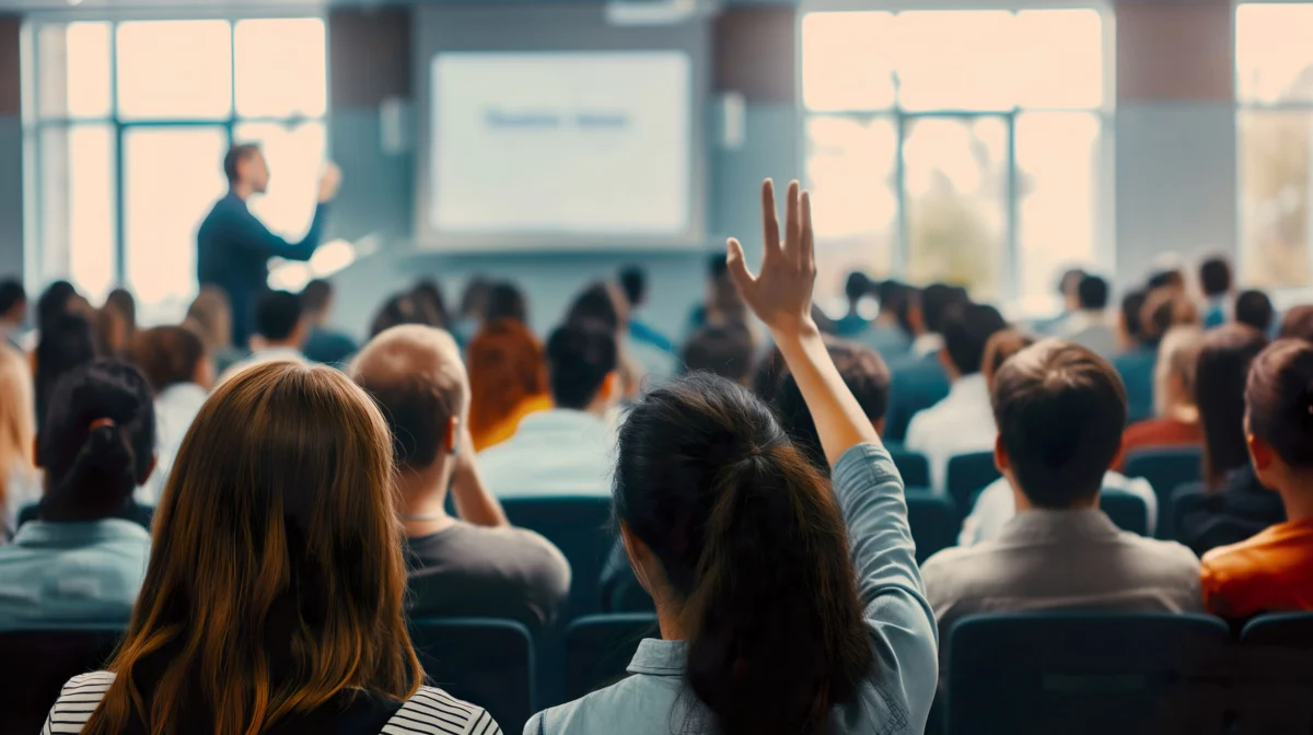 A person raises their hand in a crowded lecture hall while an instructor speaks at the front near a projected screen.