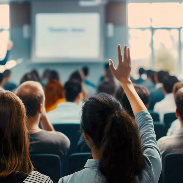 A person raises their hand in a crowded lecture hall while an instructor speaks at the front near a projected screen.