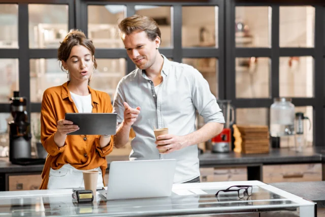 Two people standing behind a counter in a modern kitchen, looking at a tablet together. One holds a coffee cup, and a laptop and glasses are on the counter.