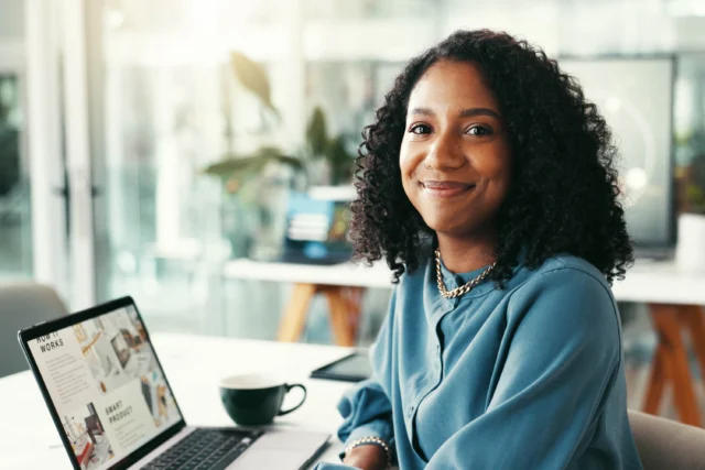 A woman with curly hair sits at a desk, smiling at the camera, with a laptop and a coffee cup in front of her in a modern office setting.