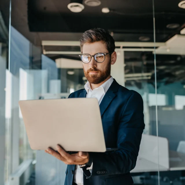 A man in a suit and glasses stands in a modern office, holding and working on a laptop.