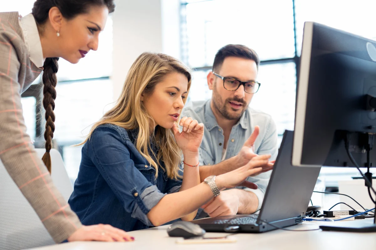 Three people working together at a desk, focused on a computer screen and discussing something, with papers and a coffee cup nearby.