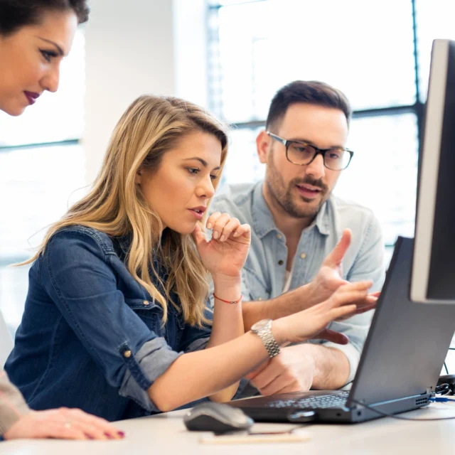 Three people working together at a desk, focused on a computer screen and discussing something, with papers and a coffee cup nearby.