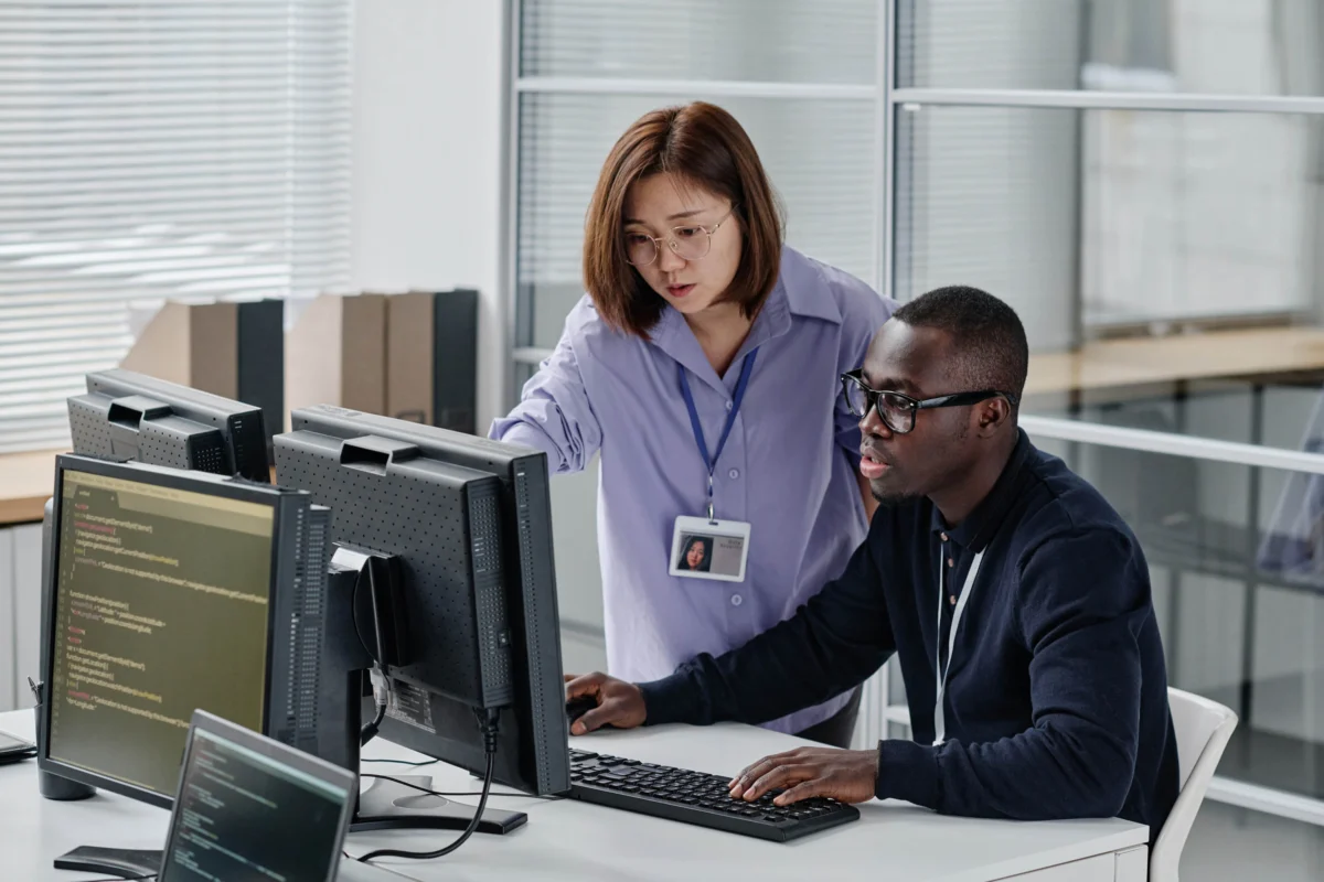 Two office workers look at computer monitors together; one is seated and typing, while the other stands and points at the screen, both focusing on the task.