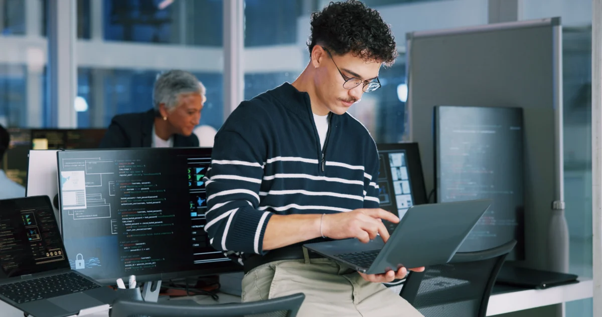 A man in glasses works on a laptop while sitting on a desk in an office, with computer monitors displaying code in the background.