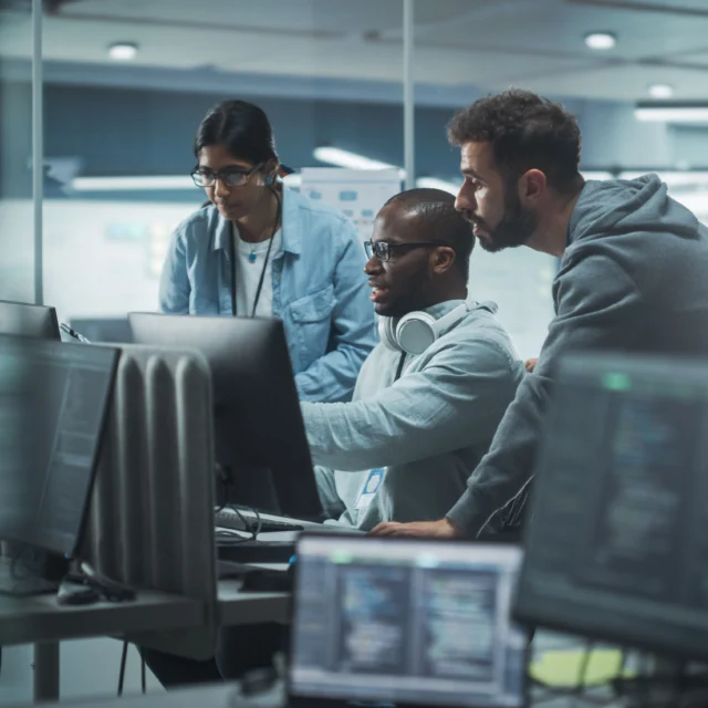 Three people in an office work together at a computer workstation with multiple monitors displaying code in a modern, glass-walled workspace.