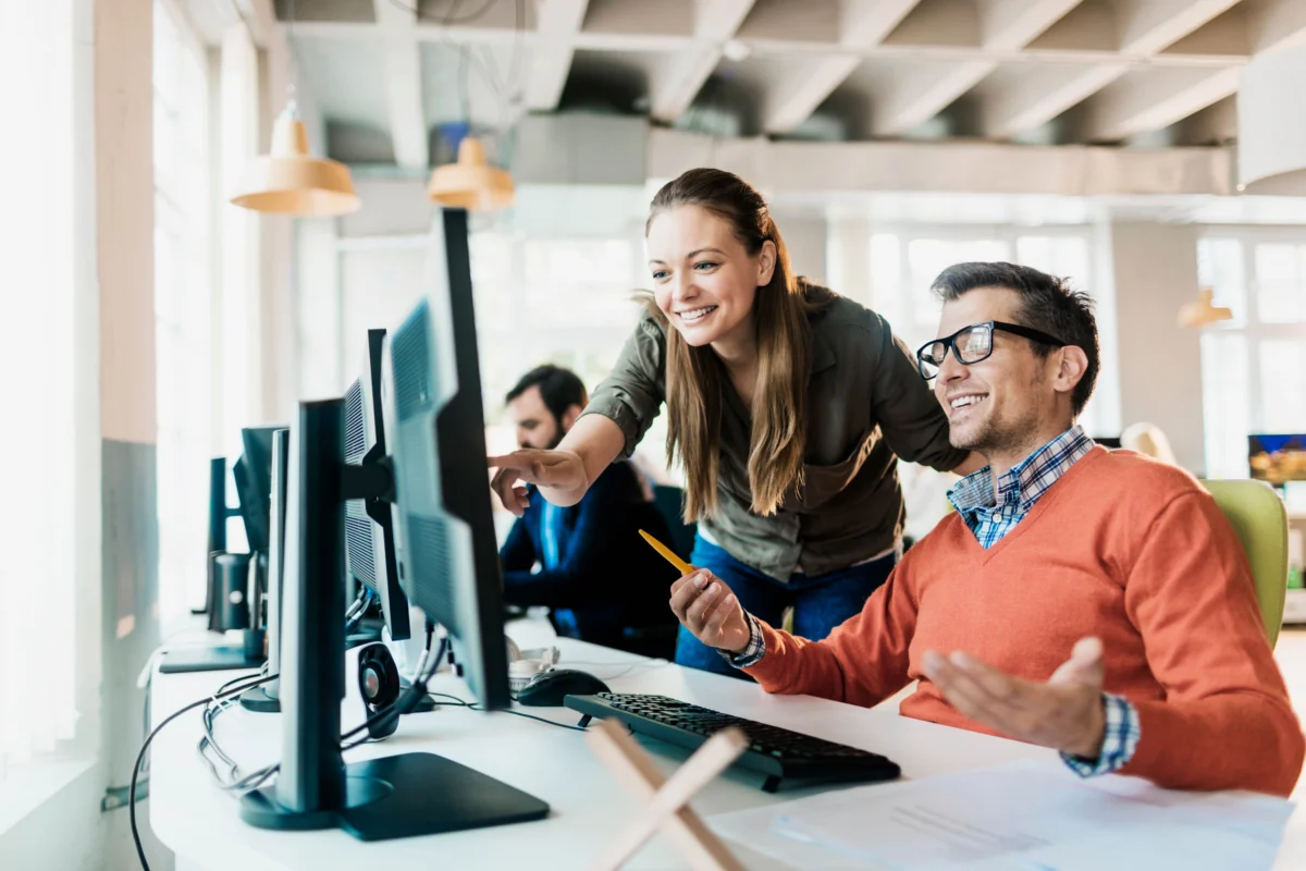Two colleagues working at computer desks; one is standing and pointing at a monitor while the other is seated, smiling and holding a pencil.
