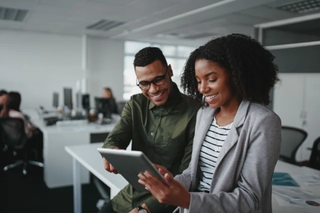 Two colleagues standing in an office, smiling and looking at a tablet together, with computer desks and chairs in the background.