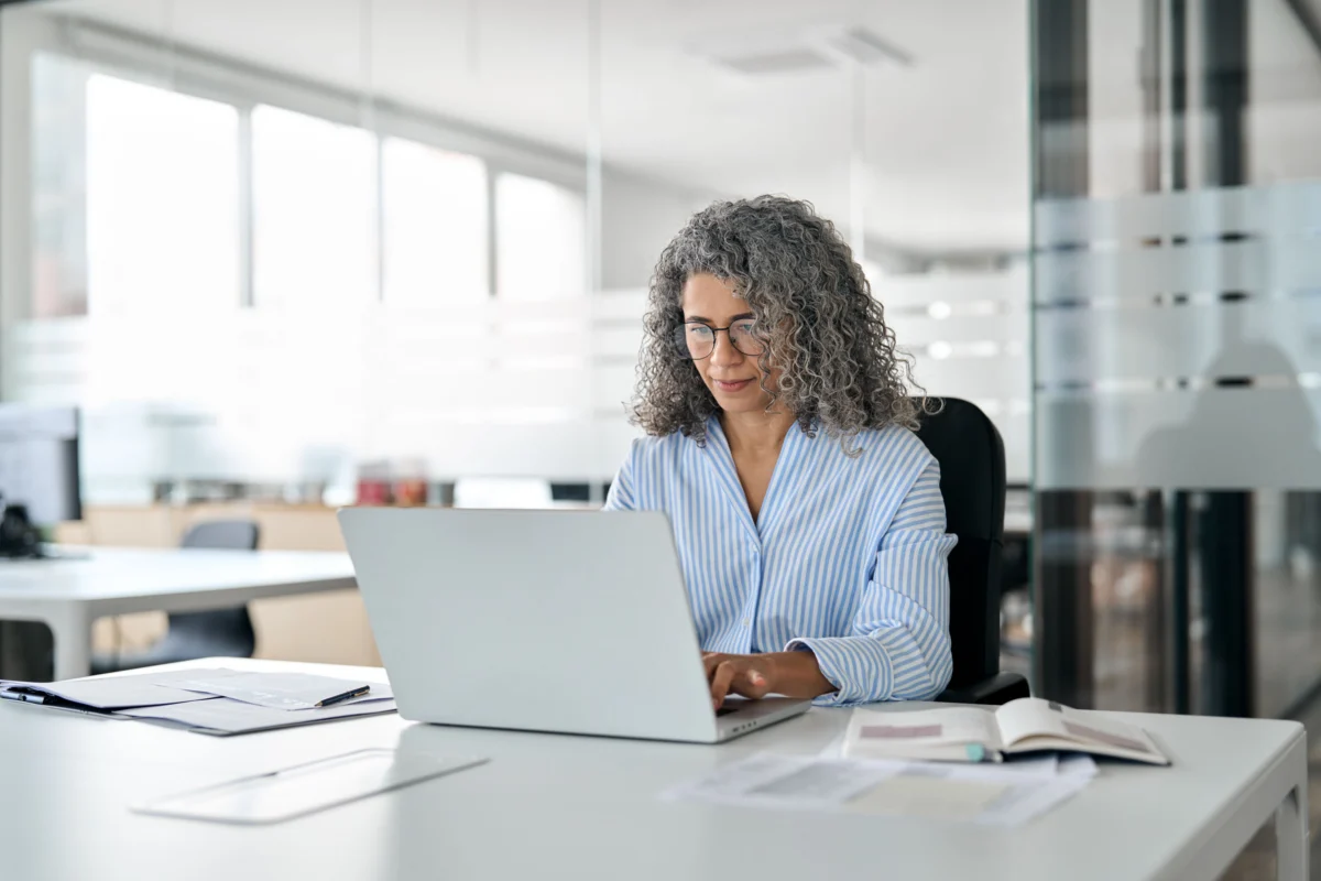A woman with curly gray hair works on a laptop at a desk in a modern office, with documents and an open notebook in front of her.