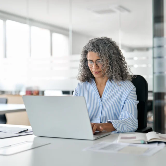 A woman with curly gray hair works on a laptop at a desk in a modern office, with documents and an open notebook in front of her.