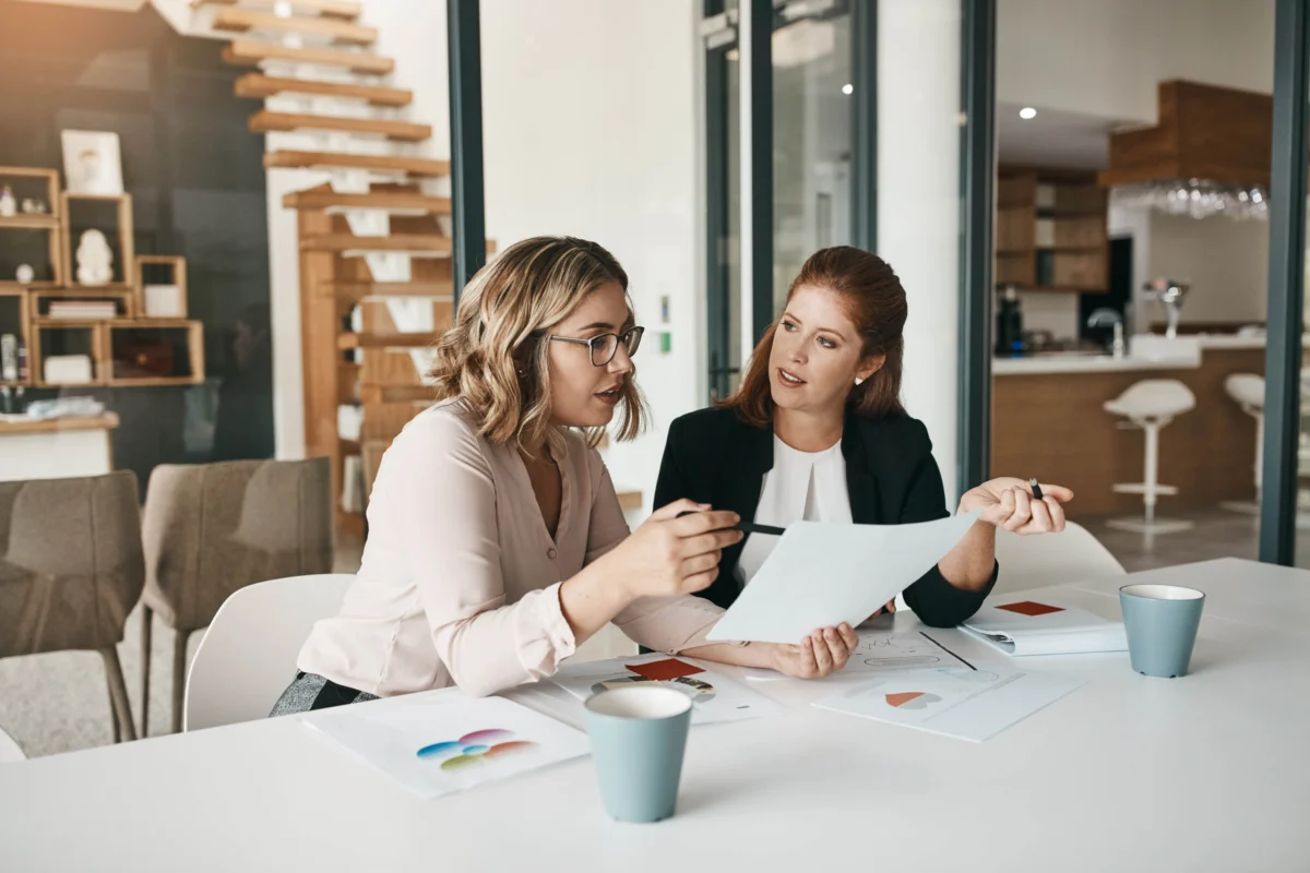 Two women sit at a table in an office, discussing documents and color charts, with coffee cups and papers in front of them.