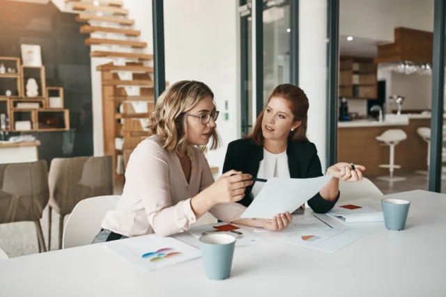 Two women sit at a table in an office, discussing documents and color charts, with coffee cups and papers in front of them.