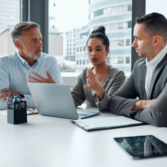 Four people sit at a desk in a modern office, having a discussion with a laptop and documents in front of them.