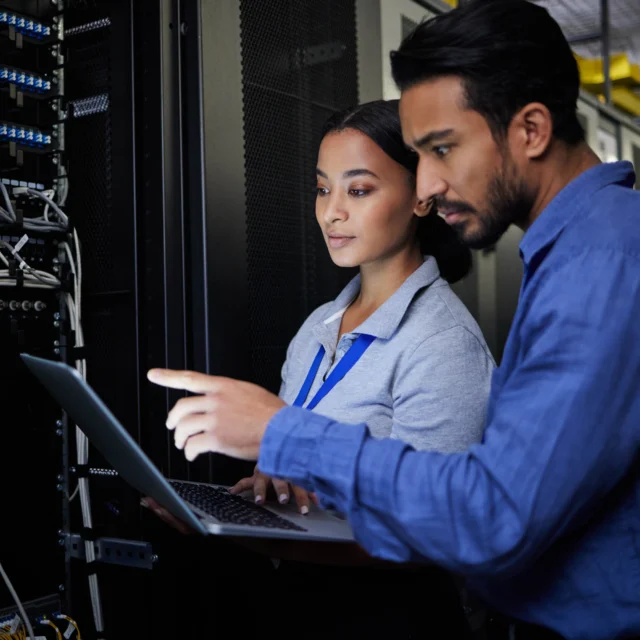 Two people stand in a server room; one holds a laptop while the other points at the screen, both focused on the computer equipment and cables around them.