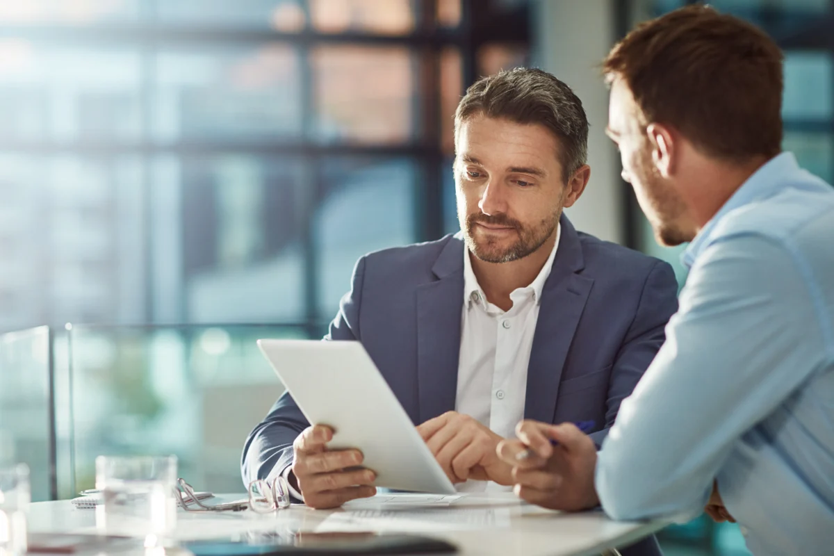 Two men in business attire sit at a table in an office, looking at a digital tablet and discussing documents.
