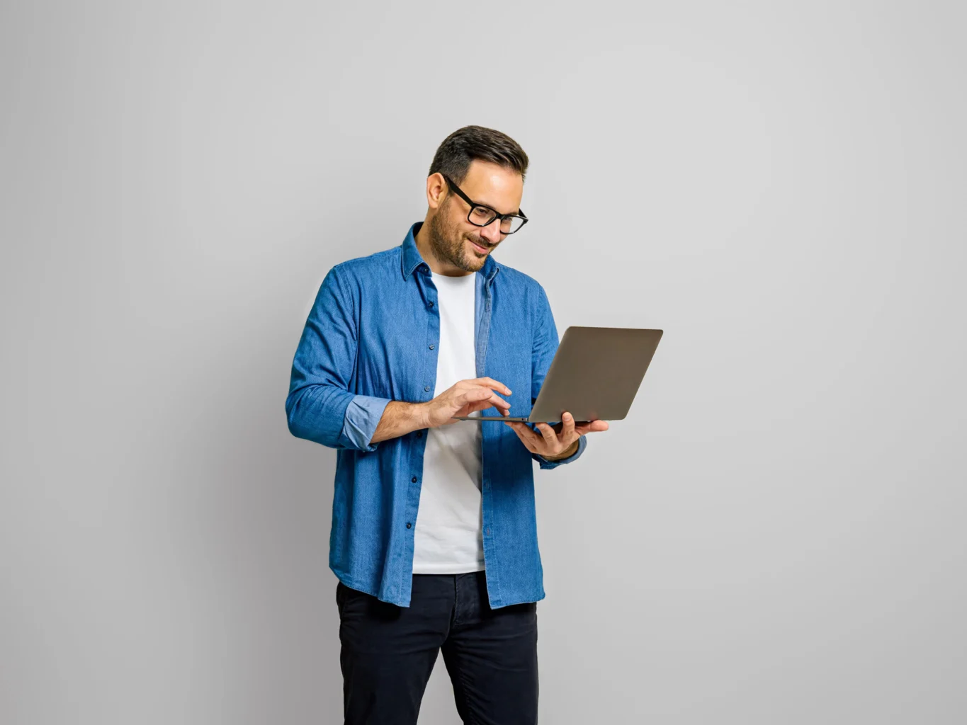 Man wearing glasses and a blue shirt stands against a plain background, smiling while using a laptop.