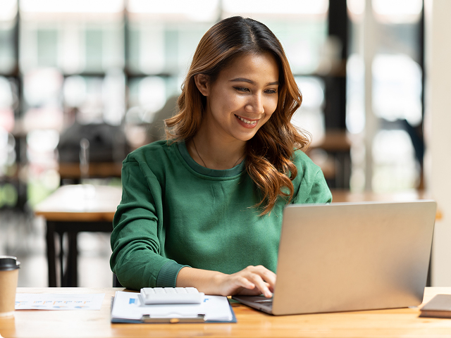 A woman in a green sweater is smiling while working on a laptop at a table with papers and a coffee cup in a bright, modern workspace.