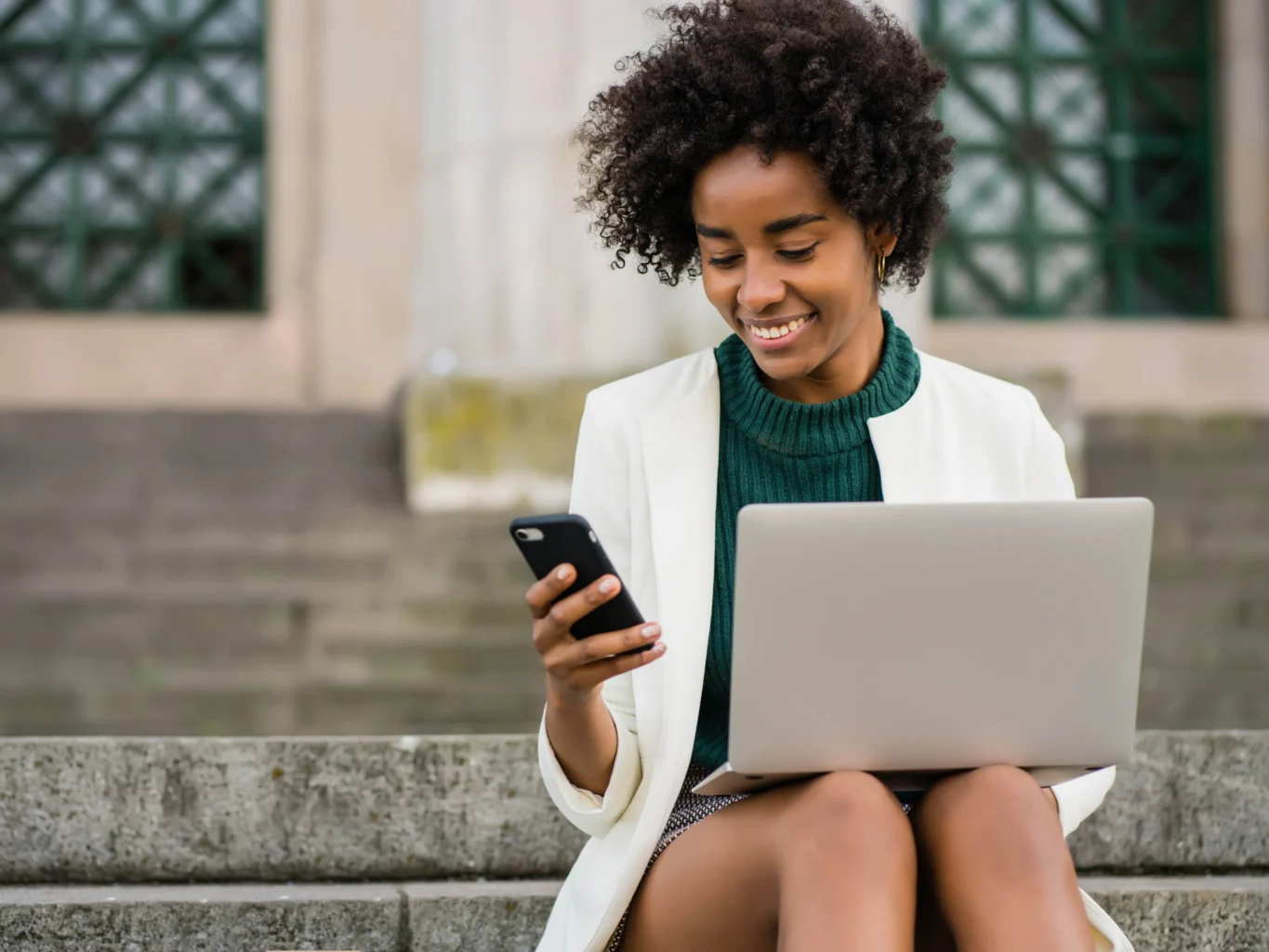 A woman sitting on outdoor steps holds a smartphone in one hand and a laptop on her lap, smiling as she looks at her phone.