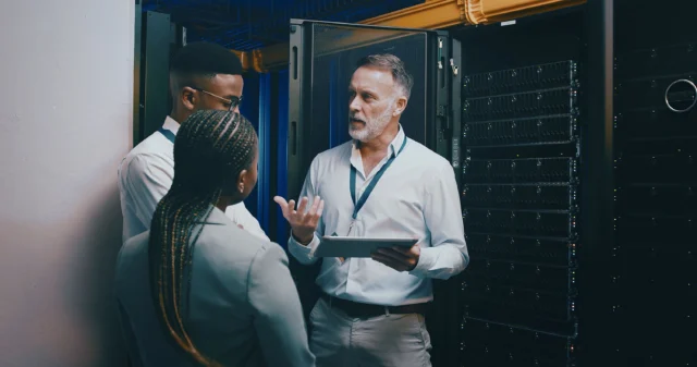 Three people stand in a server room discussing while one holds a tablet, surrounded by open server racks and network cables.