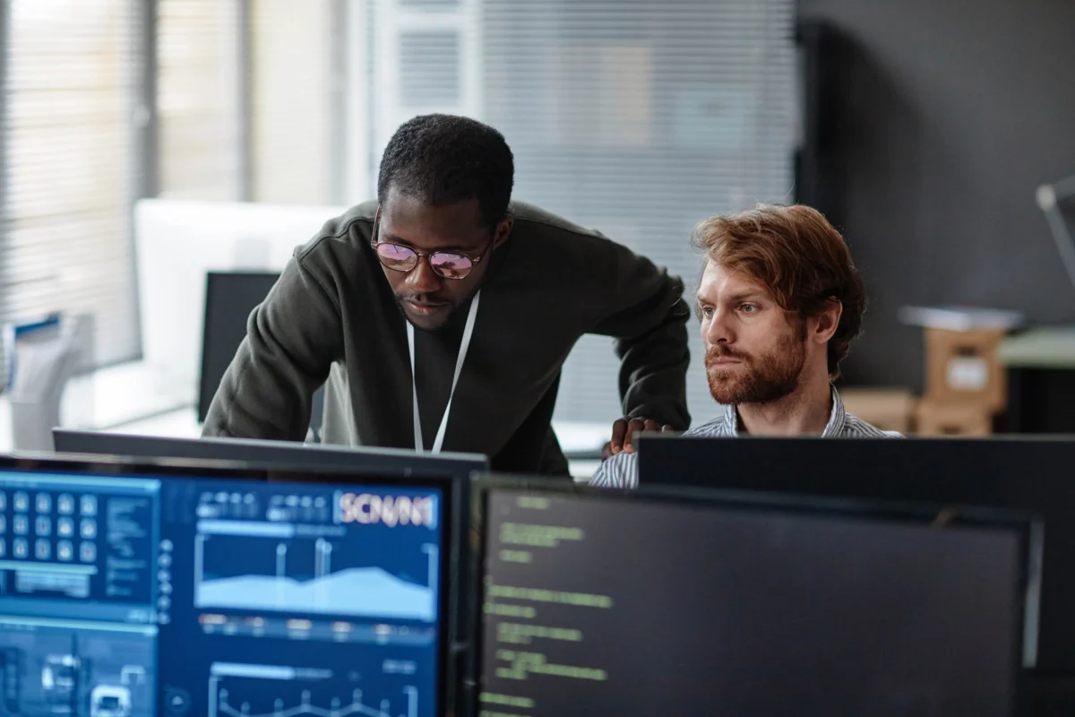 Two men look at computer screens displaying data charts and code in a modern office setting.