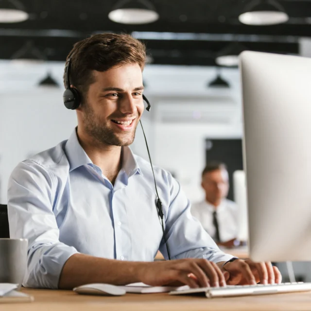 Man wearing a headset sits at a desk, typing on a computer keyboard and smiling in a modern office setting.