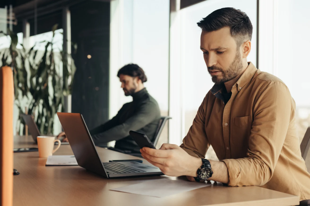 Serious male company employee checking mobile phone, using laptop, working with male colleague at open space office