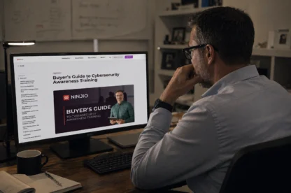 Middle-aged man with slight stubble sits at a wooden desk in a warm, lived-in office, looking thoughtfully at a desktop monitor displaying NINJIO’s Buyer’s Guide to Cybersecurity Awareness Training webpage, with shelves and soft lighting in the background.