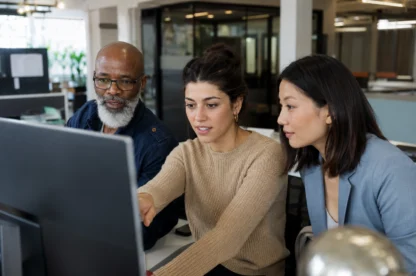 Three people sit together at an office desk, looking at a computer monitor as the woman in the middle points at the screen.